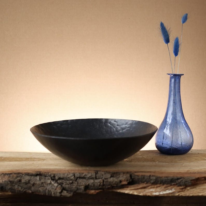 May include: A matte black bowl with a hammered texture sits next to a translucent blue glass vase containing three blue dried floral stems. The bowl has a wide, shallow circular shape, while the vase features a narrow neck and a bulbous base.