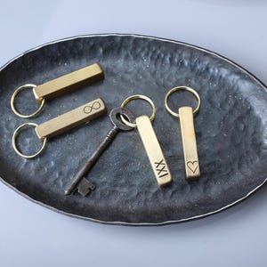 May include: A collection of keychains and a vintage key on a dark, textured oval tray. The keychains are rectangular, gold-colored, and feature engraved symbols like an infinity symbol and a heart. The tray has a metallic sheen.
