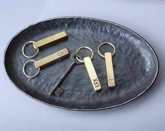 May include: A selection of keyrings and a vintage key on a dark, textured oval tray. The keyrings are rectangular, gold-coloured, and feature engraved symbols like an infinity symbol and a heart. The tray has a metallic sheen.