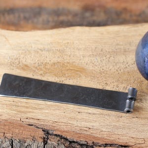 May include: A dark, rectangular metal object with a textured surface and rounded edges. It has a small, cylindrical attachment on one end. The object is set on a wooden surface, with a blue glass object in the background.