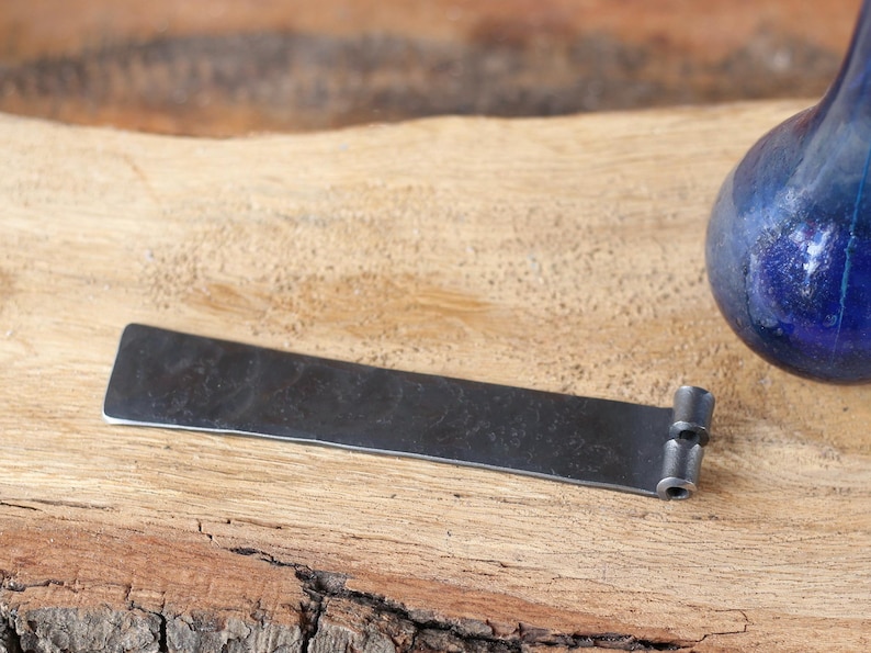 May include: A dark, rectangular metal object with a textured surface and rounded edges. It has a small, cylindrical attachment on one end. The object is set on a wooden surface, with a blue glass object in the background.