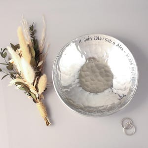 May include: A round, hammered silver-toned metal bowl featuring the engraved text 11 Juin 2016: Sab & Alex - 10 ans d&eacute;j&agrave;! alongside a small bouquet of dried beige grasses and green eucalyptus leaves, plus two thin silver-toned metal rings.