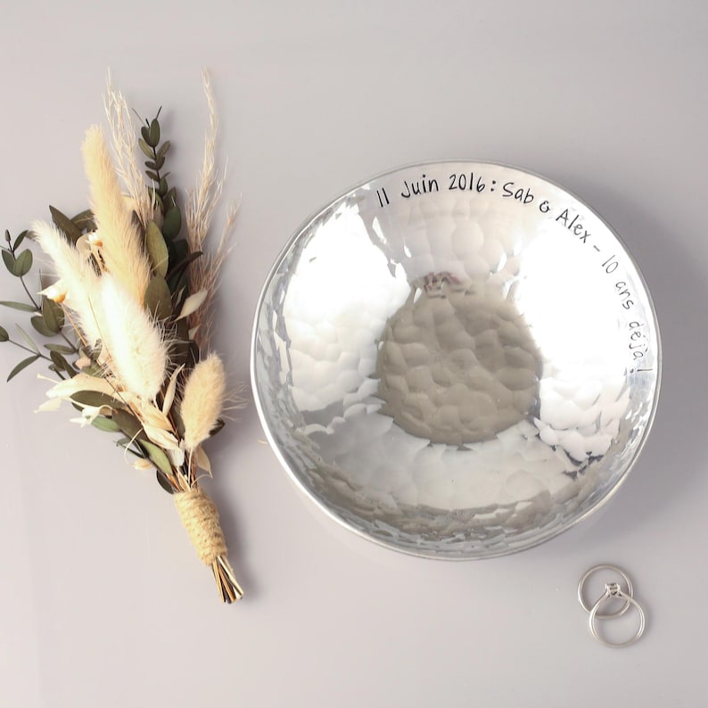 May include: A round, hammered silver-toned metal bowl featuring the engraved text 11 Juin 2016: Sab & Alex - 10 ans d&eacute;j&agrave;! alongside a small bouquet of dried beige grasses and green eucalyptus leaves, plus two thin silver-toned metal rings.