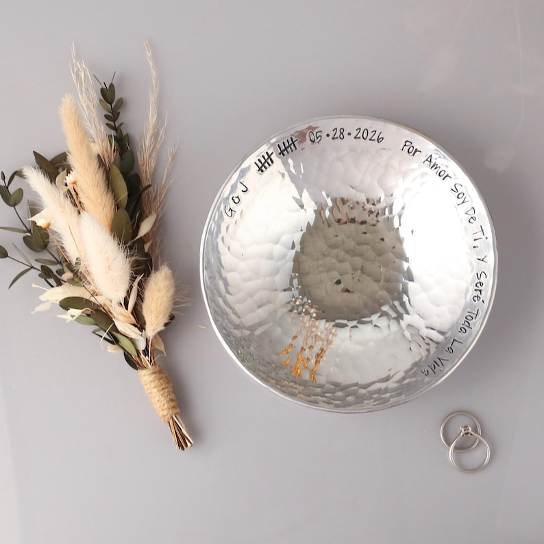 May include: A silver-toned hammered metal bowl with a circular shape, featuring engraved text that reads "G&J", "05-28-2026", and "Por Amor Soy De Ti, Y Ser&eacute; Toda La Vida". Beside the bowl is a small bouquet of dried beige grasses and two silver rings.