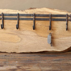 Puede incluir: Un llavero de hierro forjado con cinco ganchos, montado sobre un trozo de madera natural. El llavero de metal oscuro tiene un aspecto rústico. Un juego de llaves cuelga de uno de los ganchos. El llavero está diseñado para montaje en pared.