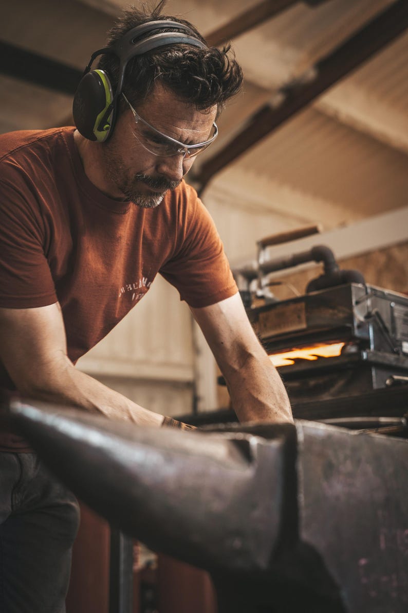 May include: A person wearing a rust-coloured t-shirt, clear safety glasses, and black ear protection works at a large metal anvil. In the background, a piece of equipment glows with orange heat, indicating a metalworking or blacksmithing environment.
