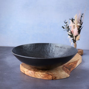 May include: A black, shallow, hammered metal bowl with a textured interior surface. The rim of the bowl is engraved with the text "Justin and Kim 1-21-2017". The bowl rests on a natural, light-coloured wooden slab with a live edge finish.