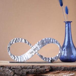 Può includere: Un simbolo dell'infinito in metallo strutturato con la scritta "Est. 2015" è esposto su una superficie di legno. Un vaso blu con steli decorativi blu è a destra. Lo sfondo è di colore beige.