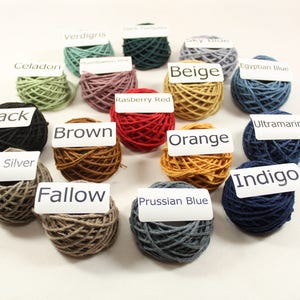 May include: A collection of yarn balls in various colors, including black, brown, orange, indigo, celadon, verdigris, raspberry red, beige, Prussian blue, ultramarine, dark silver, and fallow. Each ball has a label with the color name.