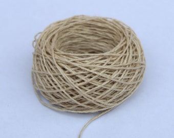 May include: A ball of natural twine, made from a natural fiber, is wound into a neat circle. The twine is a light beige color and has a rough texture. A long strand of twine is hanging from the ball.