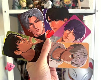 May include: A hand holding five rectangular art prints featuring anime-style male character portraits. Each print showcases a different character with varying hair colors and expressions, set against colorful backgrounds. The prints are displayed in front of a shelf with figurines.