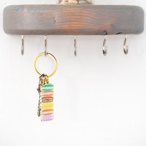 May include: A grey wooden shelf with five metal hooks and a small potted succulent plant. A gold key chain with a stack of colourful macarons hangs from one of the hooks.