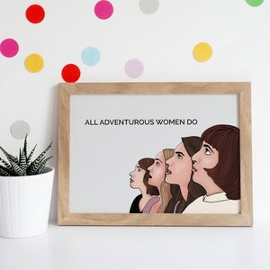 May include: A framed print with the text "ALL ADVENTUROUS WOMEN DO" and an illustration of five women looking upwards. The print is in a light wood frame. Colorful polka dots decorate the white wall behind the print.