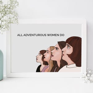 May include: A framed print with the text "ALL ADVENTUROUS WOMEN DO" above a line of five women's faces in profile. The artwork is in a white frame, with a light gray background. A vase with white flowers is in the foreground.
