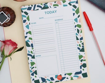 May include: A notepad with a terrazzo pattern border in shades of green, blue, and pink. The notepad has sections for 'Today', 'To Do', 'Schedule', and 'Notes'. A red pen rests on the right side. The notepad is on a light yellow surface.