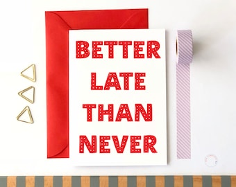 May include: A white card with red polka dot letters that read "Better Late Than Never". The card is sitting on a white surface with a red envelope, gold paper clips, and a purple and white striped washi tape.