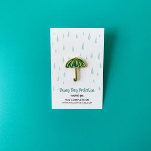 May include: Green enamel pin shaped like an umbrella with a gold outline. The pin says "Rainy Day Protection enamel pin HUE COMPLETE ME www.huecompleteme.com" on a white background with a pattern of blue raindrops.