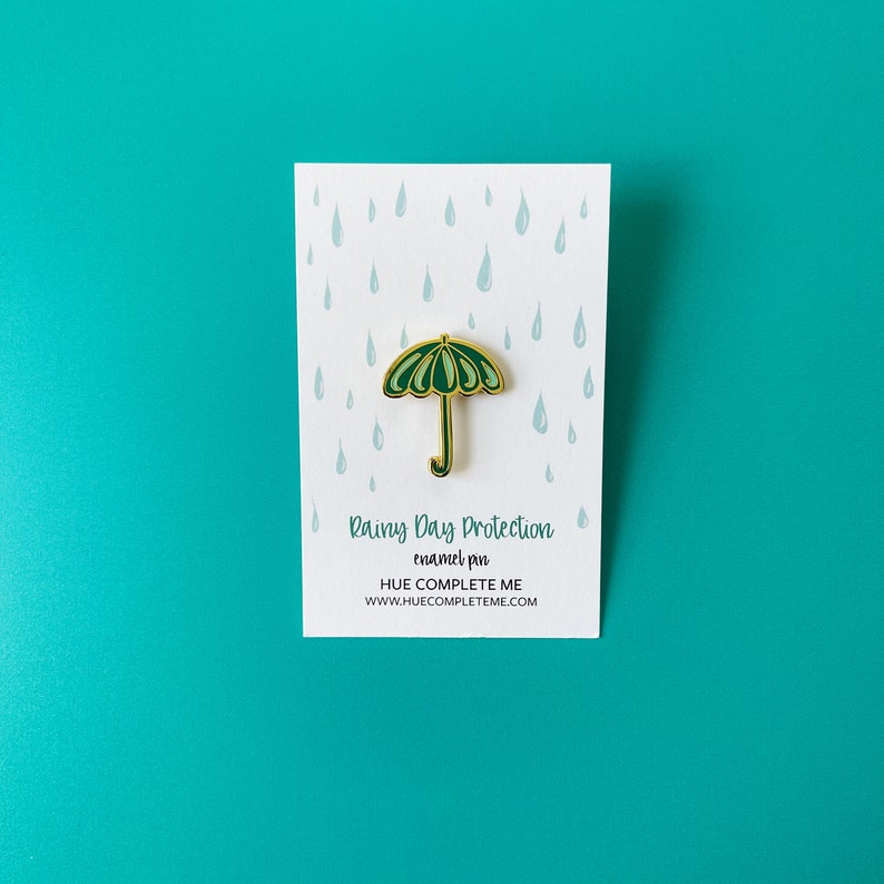 May include: Green enamel pin shaped like an umbrella with a gold outline. The pin says "Rainy Day Protection enamel pin HUE COMPLETE ME www.huecompleteme.com" on a white background with a pattern of blue raindrops.