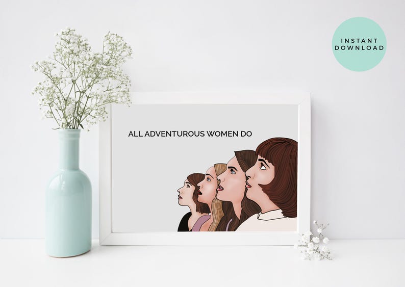 May include: A framed print with the text "ALL ADVENTUROUS WOMEN DO" above a line of five women's faces in profile. The artwork is in a white frame, with a light gray background. A vase with white flowers is in the foreground.