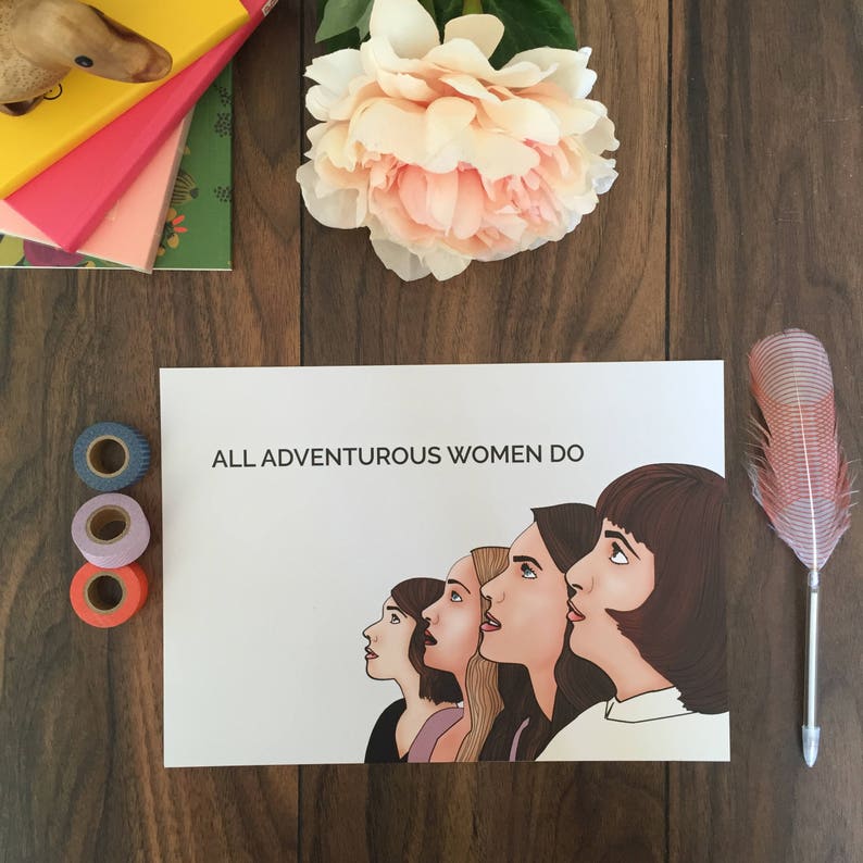 May include: A white print with the text "ALL ADVENTUROUS WOMEN DO" and an illustration of five women looking upwards. The image is on a wooden surface with a pink flower, decorative tape, and a feather pen.