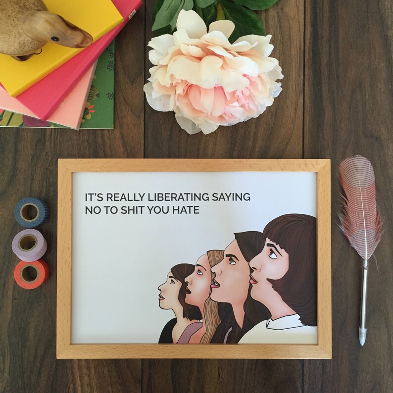 May include: A framed art print with the text "IT'S REALLY LIBERATING SAYING NO TO SHIT YOU HATE" above a line of stylized women's faces. The frame is light wood. The image is on a dark wood surface with a pink flower, a feather, and decorative tape.