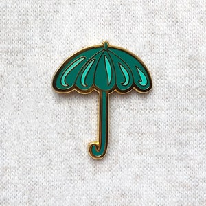 May include: A teal blue enamel pin shaped like an umbrella with a gold outline. The umbrella has a gold handle.