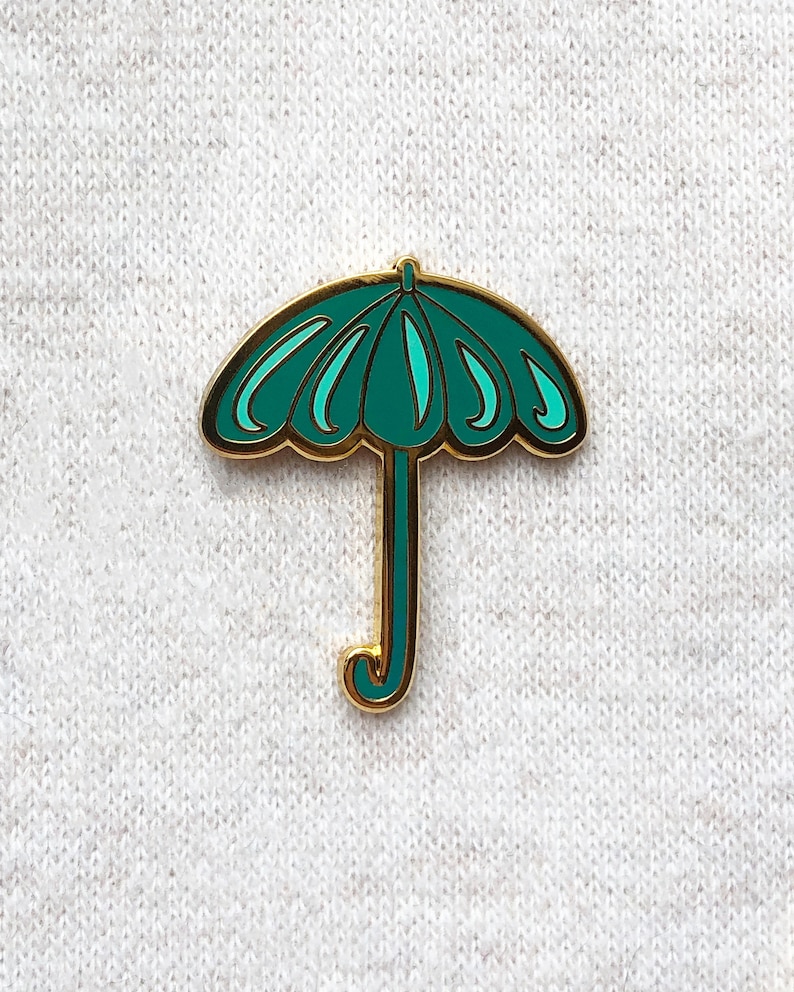 May include: A teal blue enamel pin shaped like an umbrella with a gold outline. The umbrella has a gold handle.