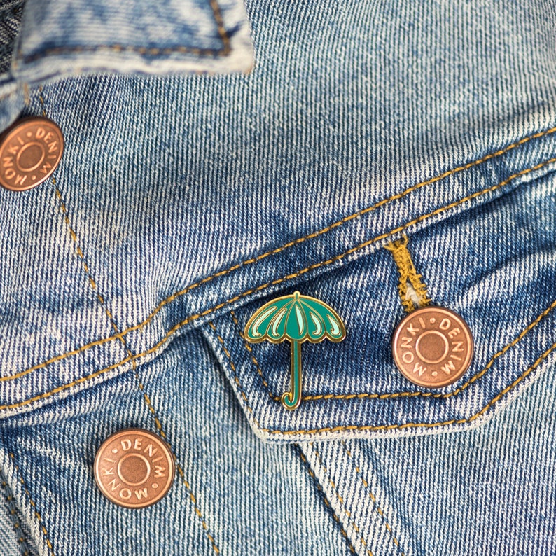 May include: A green and gold umbrella pin on a blue denim jacket with a gold button that says "Monki Denim".