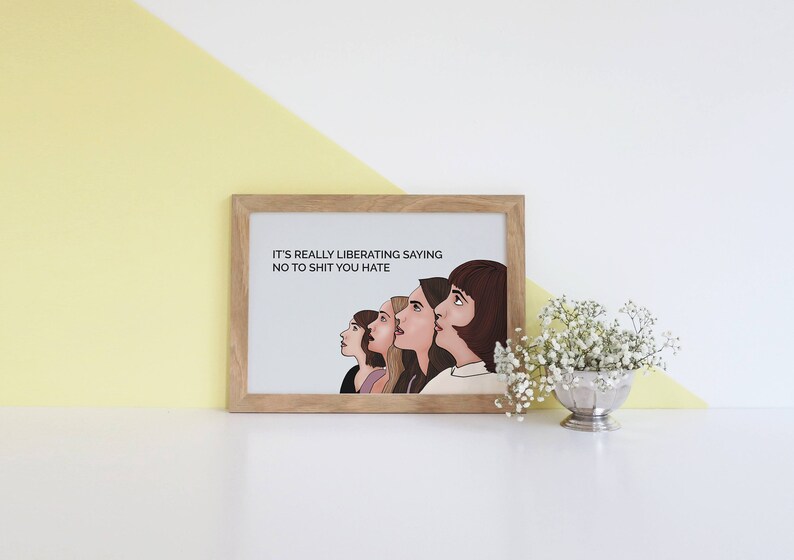 May include: A framed print in a light wood frame. The print shows five women looking upwards, with the text "IT'S REALLY LIBERATING SAYING NO TO SHIT YOU HATE". A small silver bowl of white flowers is to the right.