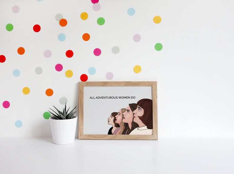 May include: A framed print with the text "ALL ADVENTUROUS WOMEN DO" and an illustration of five women looking upwards. The print is in a light wood frame. Colorful polka dots decorate the white wall behind the print.