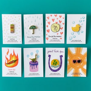 May include: A set of ten enamel pins featuring various designs, including a stack of macarons, an umbrella, a phone, a rubber duck, a chili pepper, a dinosaur on a cake, a horseshoe with a four-leaf clover, and sunglasses. Each pin is displayed on a white card with a colorful background and text that reads "HUE COMPLETE ME" and a website address.