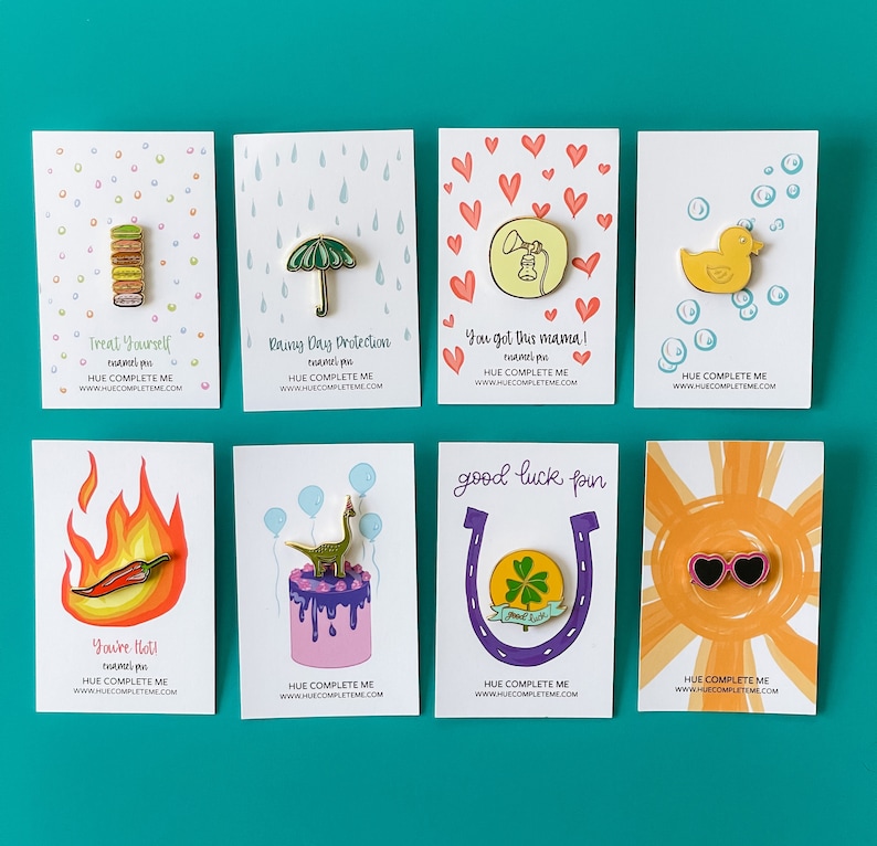 May include: A set of ten enamel pins featuring various designs, including a stack of macarons, an umbrella, a phone, a rubber duck, a chili pepper, a dinosaur on a cake, a horseshoe with a four-leaf clover, and sunglasses. Each pin is displayed on a white card with a colorful background and text that reads "HUE COMPLETE ME" and a website address.