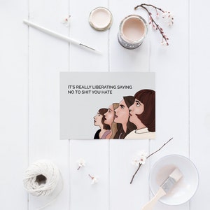 May include: A greeting card featuring the text "IT'S REALLY LIBERATING SAYING NO TO SHIT YOU HATE" and an illustration of five women. The card is on a white wooden surface, with art supplies and flowers.