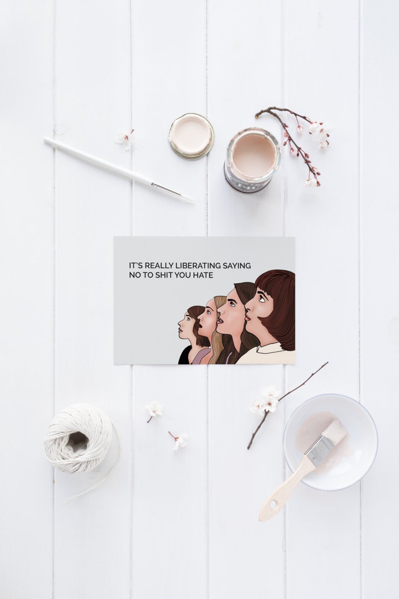 May include: A greeting card featuring the text "IT'S REALLY LIBERATING SAYING NO TO SHIT YOU HATE" and an illustration of five women. The card is on a white wooden surface, with art supplies and flowers.