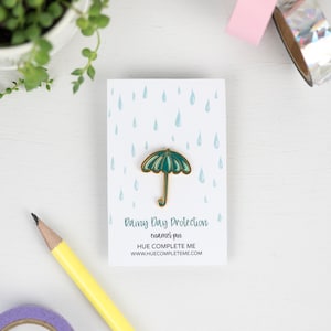 May include: Green enamel pin with a gold outline of an umbrella with raindrops in the background. The text on the card reads "Rainy Day Protection enamel pin HUE COMPLETE ME www.huecompleteme.com"