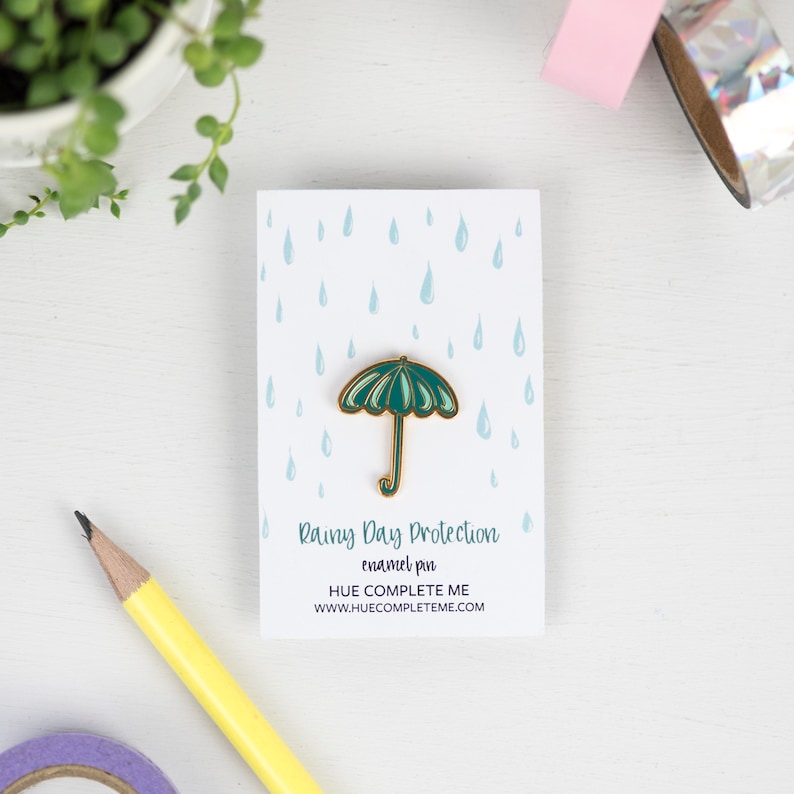 May include: Green enamel pin with a gold outline of an umbrella with raindrops in the background. The text on the card reads "Rainy Day Protection enamel pin HUE COMPLETE ME www.huecompleteme.com"