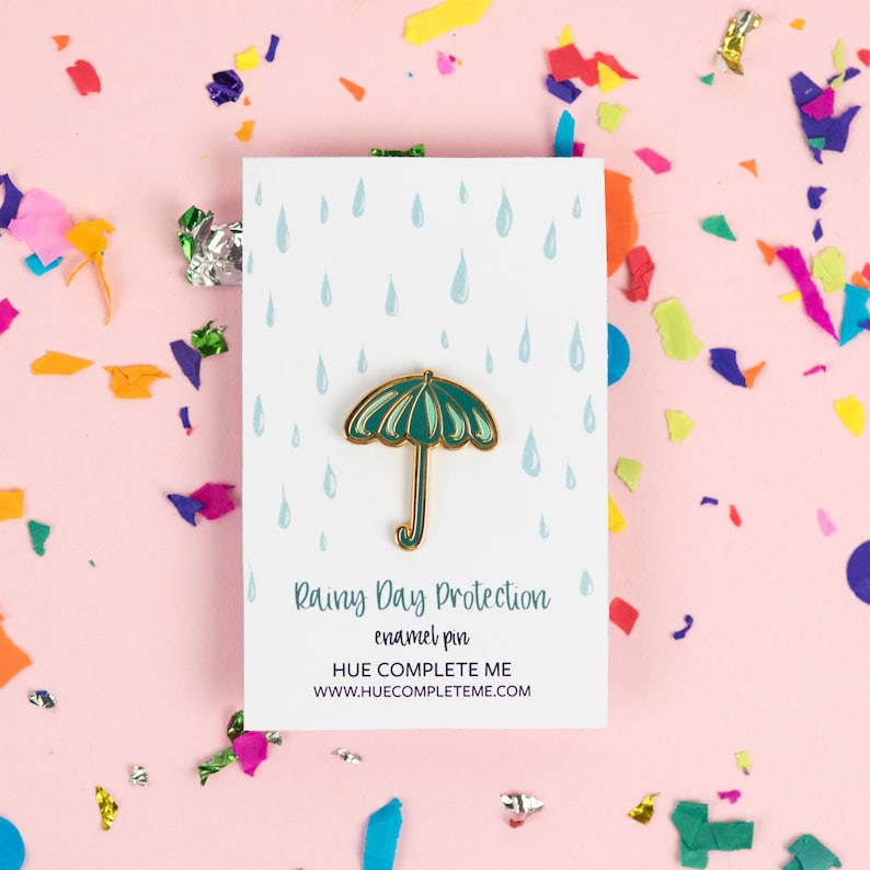 May include: A green enamel pin shaped like an umbrella with a gold handle. The pin is on a white card with a blue raindrop pattern. The card reads "Rainy Day Protection enamel pin HUE COMPLETE ME WWW.HUECOMPLETEME.COM".