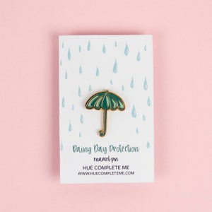 May include: Green enamel pin shaped like an umbrella with a gold outline. The pin is on a white card with a blue raindrop pattern. The card reads "Rainy Day Protection enamel pin HUE COMPLETE ME www.huecompleteme.com"