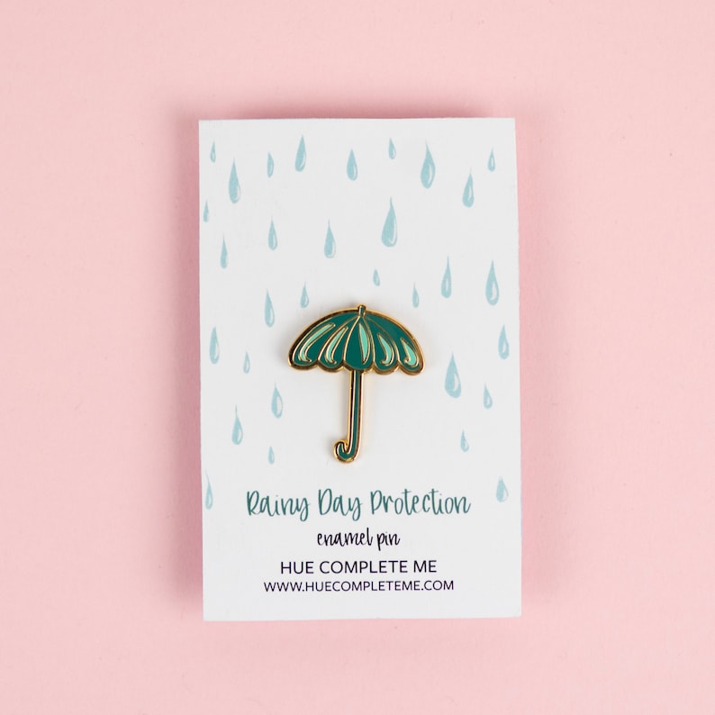 May include: Green enamel pin shaped like an umbrella with a gold outline. The pin is on a white card with a blue raindrop pattern. The card reads "Rainy Day Protection enamel pin HUE COMPLETE ME www.huecompleteme.com"