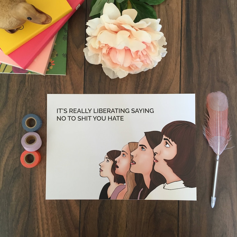 May include: A print with the text "IT'S REALLY LIBERATING SAYING NO TO SHIT YOU HATE" and an illustration of five women looking upwards. The print is on a wooden surface with decorative items like washi tape, a feather pen, and a flower.