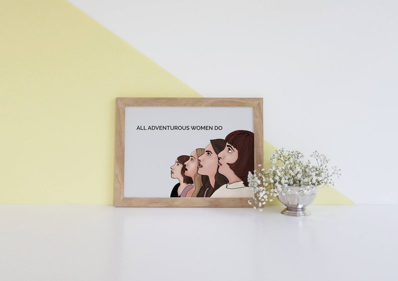 May include: A framed print with the text "ALL ADVENTUROUS WOMEN DO" above an illustration of five women looking upwards. The artwork is in a light wooden frame, set against a white and yellow background.