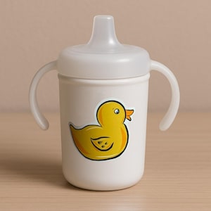 Puede incluir: Vaso para sorber blanco con dos asas y tapa blanca. Un pato amarillo de dibujos animados con pico naranja está en la parte delantera del vaso. El vaso está diseñado para niños pequeños.