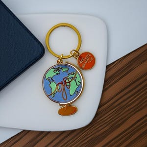Puede incluir: Un llavero dorado con un diseño de globo terráqueo con océanos azules y continentes verdes. La palabra "go" está escrita en oro sobre el globo. Un pequeño dije naranja con las palabras "true complete me" cuelga al lado.