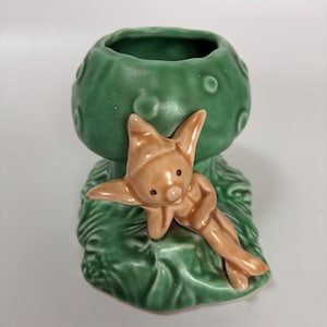 May include: A green ceramic planter shaped like a mushroom, with a small, tan-coloured fairy figure resting on the side. The fairy has pointed ears and is lying down. The planter has a textured surface.