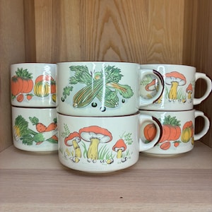 May include: Six cream-coloured ceramic mugs with brown rims, each decorated with illustrations of vegetables and mushrooms in orange, green, and yellow. The mugs are stacked on a wooden shelf, showcasing a rustic design.