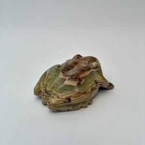 May include: A polished stone figurine carved into the shape of a frog. The sculpture features natural banding in shades of sage green, cream, and dark brown, with a smooth, glossy finish that highlights the layered mineral patterns throughout.