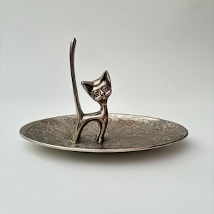 May include: A silver-toned metal ring holder featuring a stylised cat figure with an elongated tail standing upright in the centre of an oval-shaped tray. The tray surface is decorated with an intricate, embossed floral pattern throughout.