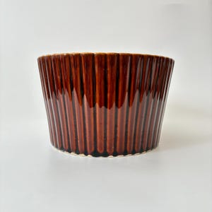May include: A deep brown ceramic bowl featuring a glossy finish and a vertical ribbed texture. The bowl has a wide, open top and a tapered base, with the glaze showing subtle variations in colour intensity along the vertical ridges.