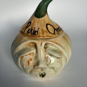 May include: A ceramic jar in the shape of an onion, featuring a face design. The jar is beige with green and brown details. The lid has a green stem and the words "Pickled Onions" are written in black script.