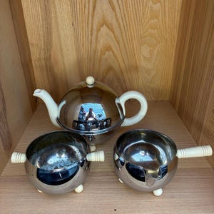 May include: A vintage tea set with a reflective chrome finish. The set includes a teapot with a white handle and spout, a sugar bowl, and a creamer. Each piece has small, white feet and handles.
